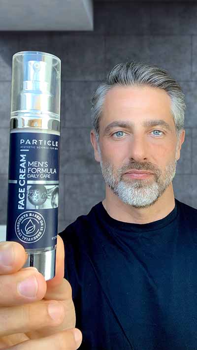 A close-up of a bottle of Particle Men's Formula Face Cream held by a man.