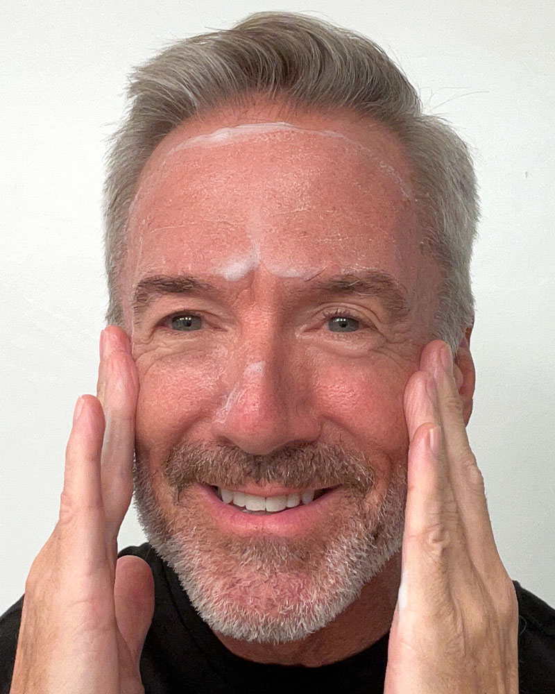 Middle-aged man applying cream to his face, smiling.