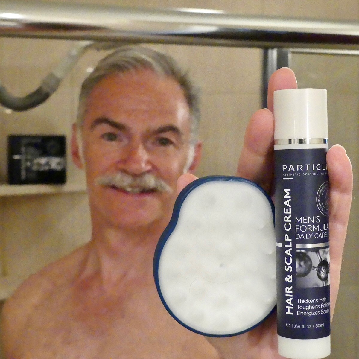 A shirtless man holds a Particle hair and scalp cream and a scalp massager in a bathroom.