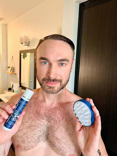 A shirtless man holding a Particle hair product and a scalp brush in the room.