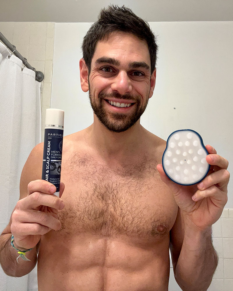  Smiling man in a bathroom, holding a bottle of Hair & Scalp Cream and a scalp brush.