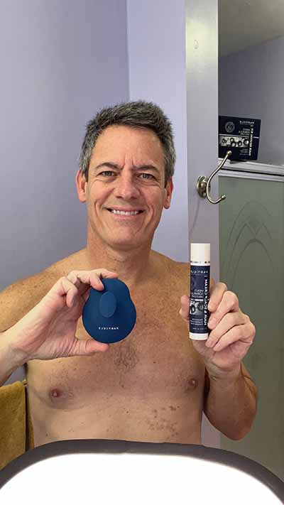 Shirtless man smiling, holding blue device and Particle hair product in bathroom.