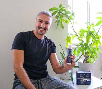 Smiling man holding a Particle product bottle, seating near potted plants.