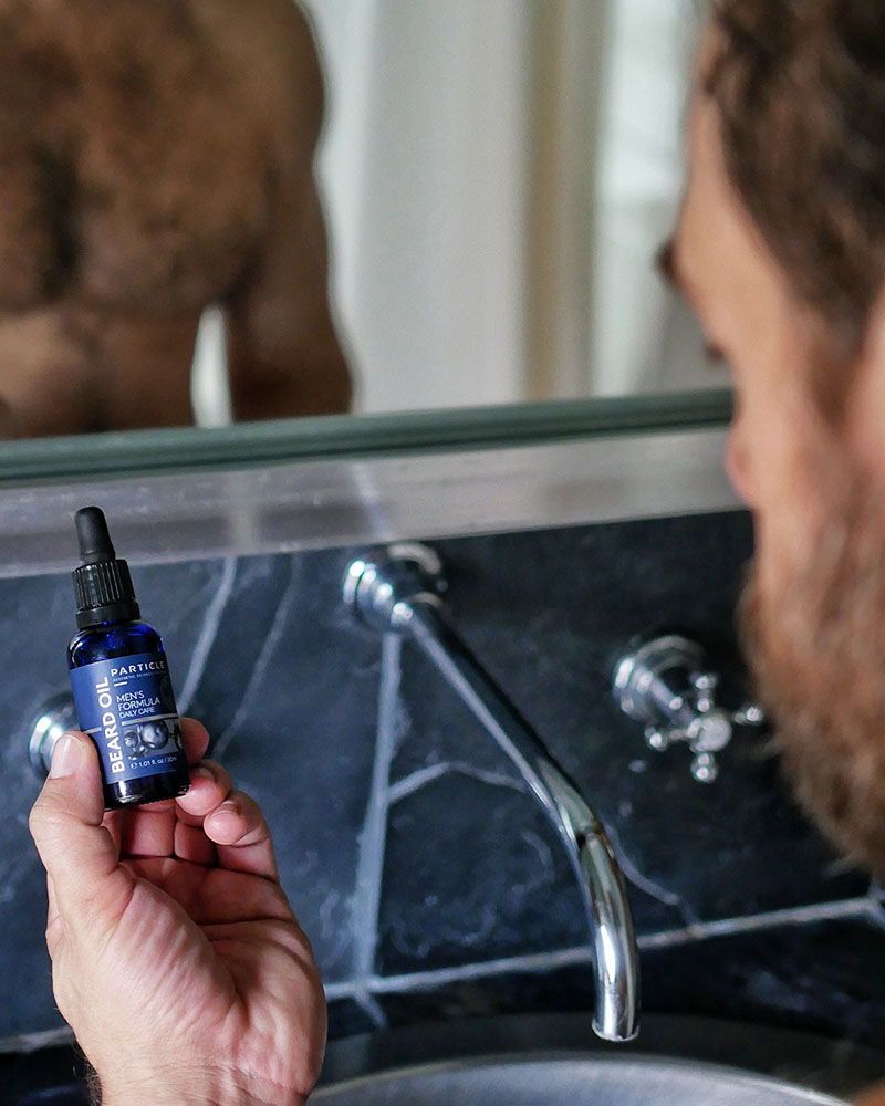 Hand holding Particle Beard Oil dropper bottle by bathroom sink.