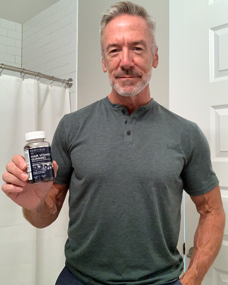 Man holding a clear bottle of Particle Hair Vitamin Gummies with white cap and dark blue label.