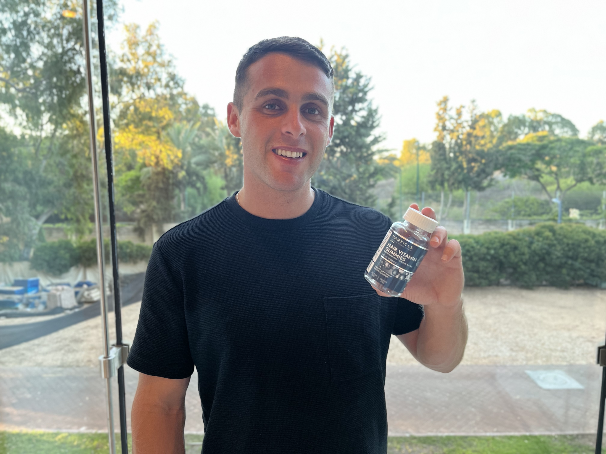 A young man holds a bottle labeled Particle Hair Vitamin Gummies.