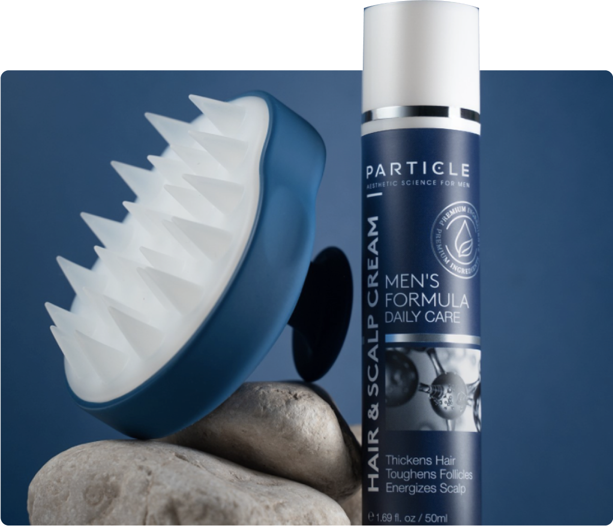 Particle Hair and scalp cream bottle stands beside a spiked scalp massager on rocks.