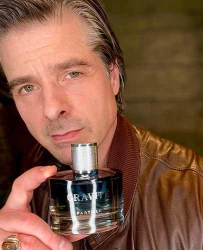 Man wearing a brown jacket holds a perfume bottle labeled Gravité Particle.